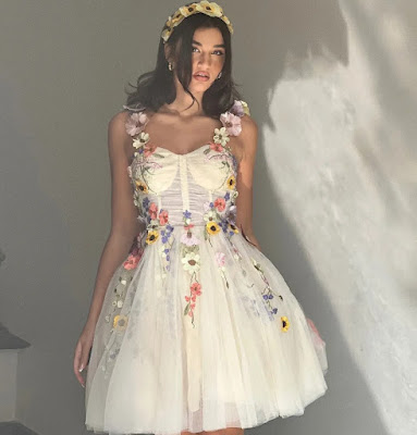 A young woman posing in a cream-colored, tulle mini dress featuring vibrant 3D floral appliqués and embroidery. She is wearing a matching floral headband that complements the whimsical and romantic aesthetic, perfect for a creative or informal garden wedding.