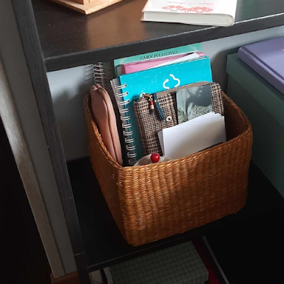 Cesta de palha colocada numa prateleira de escritório, contendo cadernos, blocos, estojos e documentos organizados.