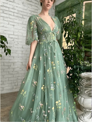 A woman wearing a floor-length, sage green tulle dress with a deep V-neckline and puff sleeves. The gown is adorned with delicate colorful floral embroidery, featuring tiny yellow and pink blossoms. The dress has a corset-inspired bodice with ribbon ties, creating an ethereal and fairy-tale aesthetic perfect for a formal garden wedding or a woodland ceremony.