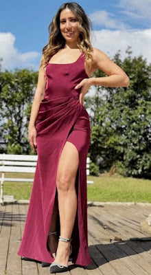 Foto de corpo inteiro de uma mulher posando em um deck de madeira ao ar livre. Ela usa um vestido longo na cor vermelho borgonha, com decote reto, drapeado na cintura e uma fenda lateral profunda. Ela calça sapatos de salto alto pretos com detalhes brilhantes no tornozelo e na ponta.