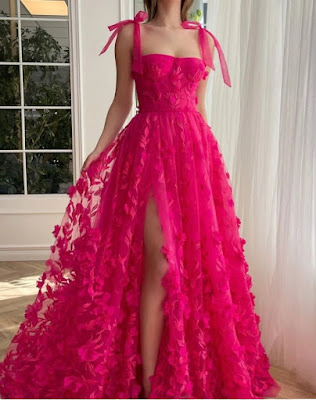 A full-length photo of a woman in a dramatic, bright fuchsia pink gown. The dress is strapless with a corset-style bodice and shoulder tie-straps. It is covered in intricate 3D floral appliqués and embroidery. A high slit reveals her leg. The aesthetic is bold, glamorous, and eye-catching.