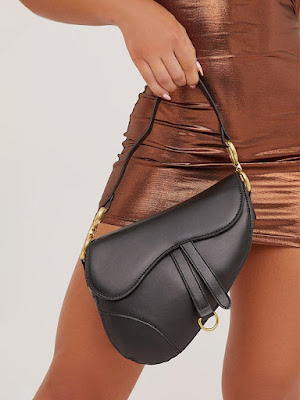 Close-up de uma pessoa a segurar uma mala de ombro preta em formato de sela (saddle bag). A mala possui detalhes metálicos dourados nas alças e tiras decorativas na frente. O look é composto por um vestido curto metalizado num tom bronze brilhante.