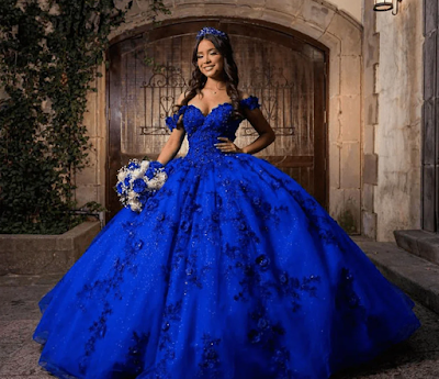 Uma imagem deslumbrante de uma jovem posando para sua Quinceañera. Ela veste um opulento vestido de baile azul royal com um corpete sem alças e uma saia volumosa, cobertos por intrincados apliques florais e glitter cintilante. Ela usa uma tiara de cristal reluzente e brincos combinando, e segura um buquê de mixed rosas brancas e azuis. O cenário é um pátio de pedra rústico com grandes portas de madeira arqueadas, iluminado por lanternas de parede quentes e adornado com trepadeiras verdes.