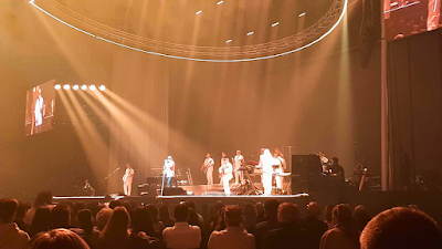 Fotografia captada da plateia, mostrando o palco dourado de um grande concerto na Super Bock Arena. No palco, iluminado por fortes focos de luz amarela, estão o cantor Pedro Abrunhosa e a sua banda, todos vestidos de branco. Um grande anel de iluminação oval está suspenso sobre o palco. Vê-se um ecrã LED gigante à esquerda projetando um grande plano de Pedro Abrunhosa. O público está sentado em primeiro plano, observando o espetáculo.