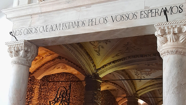 Entrada da Capela dos Ossos em Évora com a inscrição 'Nós ossos que aqui estamos pelos vossos esperamos', rodeada por pilares e paredes com ossadas humanas.