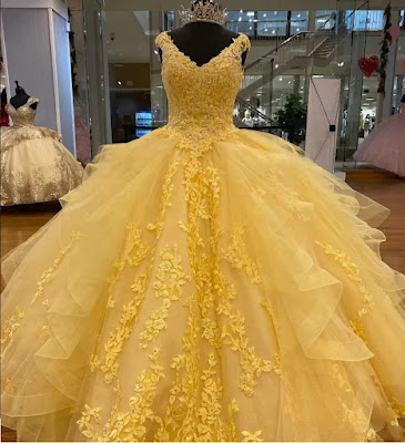 Uma imagem deslumbrante de um vestido de Quinceañera em um tom radiante de amarelo sol, exibido em um manequim em um elegante salão de baile. O vestido apresenta um corpete sem alças com intrincados bordados dourados e apliques florais cintilantes. A volumosa saia de baile é feita de camadas de tule suave e é acentuada por babados estruturados e apliques dourados verticais que descem em cascata. O manequim usa uma coroa de cristal cintilante. O ambiente inclui painéis de parede dourados, espelhos altos que refletem o ambiente e grandes árvores de cerejeira artificiais com flores rosa suave, criando um contraste dramático e sofisticado. A luz quente do lustre ilumina a cena.