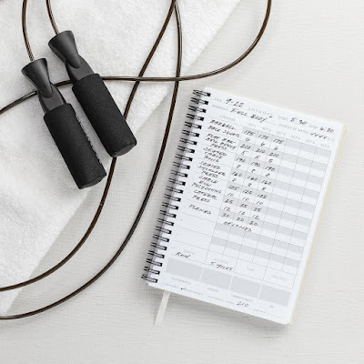 Corda de saltar e caderno de registo de treino com exercícios anotados, representando o planeamento e a criação de uma rotina de atividade física.