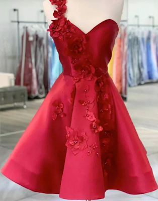 A vibrant crimson red cocktail dress displayed on a white mannequin. The dress features a unique asymmetrical design with a single shoulder strap and a sweetheart neckline. It is heavily embellished with intricate 3D floral appliqués and matching red beads that cascade from the shoulder, down the bodice, and onto the voluminous A-line skirt. The fabric has a rich, satin-like sheen, and the background shows a blurred interior of a clothing boutique with various dresses on racks.