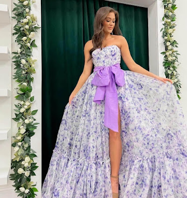 A young woman posing in an elegant, strapless ball gown featuring a white fabric printed with delicate lavender-purple flowers. A large, solid lavender bow sash is cinched at the waist, and a dramatic high slit reveals her leg and nude heel. She stands against a backdrop of deep green curtains framed by beautiful floral garlands of white roses and greenery, showcasing a stunning, romantic look for a garden ceremony.