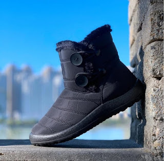 O essencial para enfrentar o frio com estilo Bota de inverno preta com interior de pelo e fecho com botões, pousada num muro de pedra com fundo urbano e céu azul, perfeita para proteger do frio.