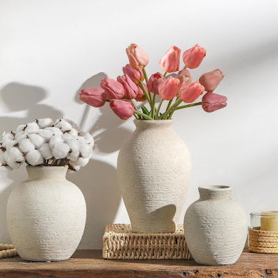 Conjunto de jarras em cerâmica clara com tulipas cor-de-rosa e flores de algodão, sobre móvel de madeira, criando uma decoração primaveril luminosa, natural e acolhedora.