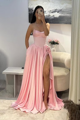 Uma jovem tira uma selfie ao espelho vestindo um longo vestido de festa rosa claro acetinado. O vestido tem um corpete justo estilo espartilho sem alças, com decote ligeiramente drapeado. A saia é volumosa e fluida, apresentando uma fenda lateral profunda decorada com aplicações de flores de tecido na anca e bolsos discretos.