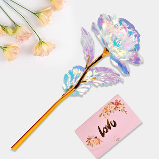 Uma rosa decorativa com pétalas de efeito holográfico iridescente e caule dourado, acompanhada por um envelope rosa com a palavra "Love" escrita em dourado e flores secas ao fundo.