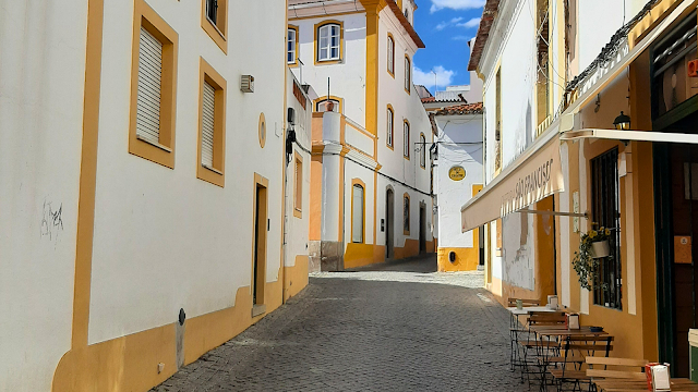 Rua estreita e pitoresca de Évora, com casas brancas e detalhes em amarelo, café com esplanada e céu azul ao fundo.