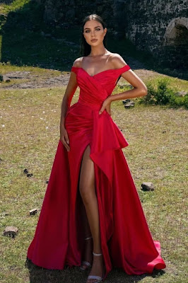 Uma mulher posa num campo com um longo vestido de festa vermelho vibrante em cetim. O modelo tem decote ombro a ombro drapeado, uma fenda profunda na perna esquerda que revela sandálias prateadas de salto alto, e uma sobressaia volumosa na anca. Ao fundo, veem-se ruínas de pedra.