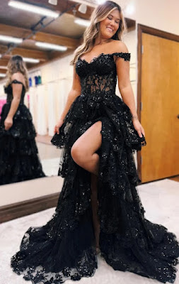 Uma mulher jovem e sorridente com cabelo castanho ondulado está de pé num salão de noivas, posando para a câmara enquanto veste um luxuoso vestido de baile preto. O vestido tem um decote ombro a ombro adornado com renda e lantejoulas, um corpete de estilo corset em renda preta e uma saia de tule e renda em camadas com uma fenda alta que revela a sua perna esquerda e uma tatuagem no tornozelo. Ela está de pé sobre um tapete branco com o seu pé esquerdo apoiado numa cadeira oculta para mostrar a fenda do vestido. Ao fundo, vê-se um espelho grande que reflete a parte de trás do vestido e a loja com mais vestidos de festa e de noiva pendurados em cabides, e um teto alto com vigas expostas e luzes de pista. O chão é de azulejos claros.
