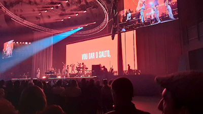 Vista de um grande palco de concerto iluminado em tons de vermelho e rosa. No centro do palco, um ecrã gigante exibe a frase "VOU DAR O SALTO," em letras brancas. A banda e o artista estão posicionados à frente do ecrã. Vários focos de luz e estruturas circulares de iluminação estão visíveis no teto da arena. Em primeiro plano, as silhuetas do público observam o espetáculo.