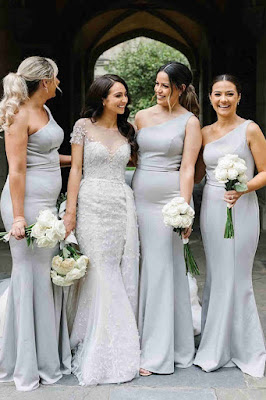 Noiva ao centro, vestida com um vestido de noiva rendado e estruturado, rodeada por três damas de honor que vestem vestidos longos iguais, de um ombro só, na cor cinza claro. Todas seguram bouquets de rosas brancas e sorriem num ambiente arquitetónico clássico.