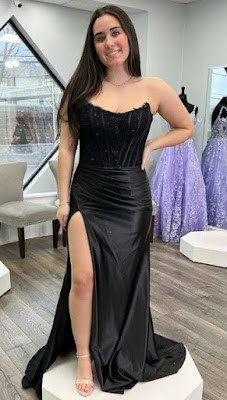 Uma mulher jovem de cabelos longos e escuros sorri enquanto posa com um vestido de festa preto comprido. O vestido destaca-se pelo contraste de materiais: o topo é um corpete justo, sem alças (tomara-que-caia), com detalhes bordados e transparências subtis, enquanto a saia é feita de um cetim preto brilhante, apresentando um drapeado na anca e uma fenda alta na perna direita. Ela está em cima de uma plataforma branca num salão de festas; ao fundo, veem-se poltronas cinzentas, uma janela ampla e outros vestidos de gala em tons de lilás expostos em manequins. O chão é de madeira clara.