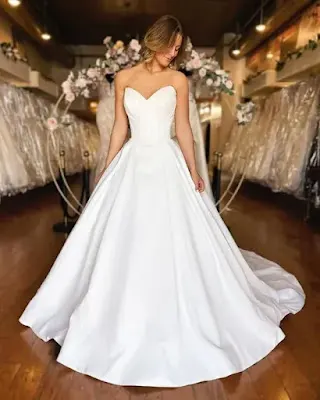 Timeless Strapless Satin Ball Gown Wedding Dress A bride standing in a bridal boutique wearing a classic, minimalist strapless ball gown wedding dress with a structured sweetheart bodice and a full satin skirt with a train.