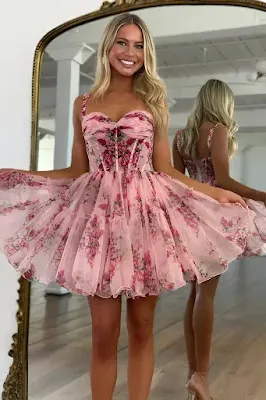 A smiling young woman with blonde hair poses in front of a mirror wearing a pink, floral-print mini dress with thin straps, a structured corset-style bodice, and a voluminous, layered tulle skirt.