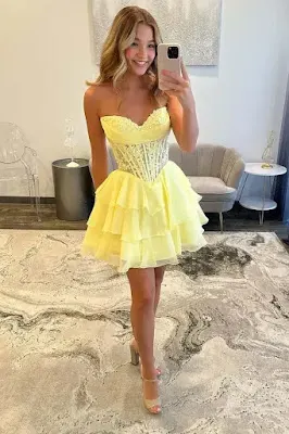A young woman with wavy blonde hair smiles while posing for a mirror selfie, holding a silver smartphone. She is wearing a strapless, short yellow dress featuring a structured corset bodice and a ruffled, layered skirt. She completes the look with clear high-heeled sandals. The reflection shows a dress boutique with a grey and white abstract-patterned rug, a transparent acrylic chair, a grey tufted sofa, and small side tables with lamps. The lighting is soft and bright.