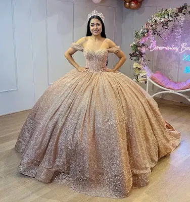 A young woman with long dark hair stands smiling, wearing a spectacular, full-skirted rose gold quinceañera ball gown with a sparkling glitter finish and off-the-shoulder sleeves. She also wears a silver tiara. The background features a floral arch and neon sign.