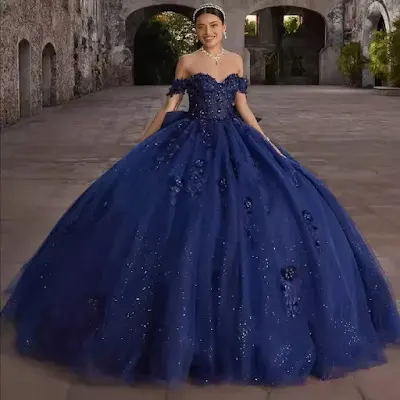 A young woman stands smiling, wearing a magnificent royal blue quinceañera ball gown. The dress features an off-the-shoulder sweetheart neckline, a corset-style bodice covered in intricate 3D floral appliqués and beading, and a voluminous, glittery tulle skirt. She is also wearing a tiara and necklace, set against the background of an old stone courtyard with archways.