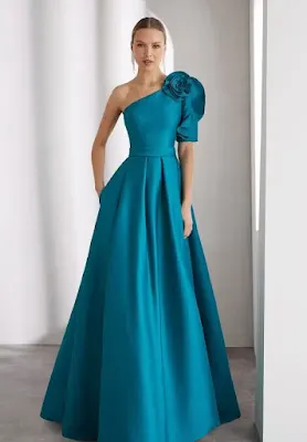 Elegant Teal One-Shoulder Evening Gown with Floral Detail A model poses in a neutral studio setting, wearing an elegant, floor-length teal evening gown. The dress features a one-shoulder neckline adorned with a large fabric flower, a cinched waist with pleating details, and a full, voluminous skirt with side pockets.