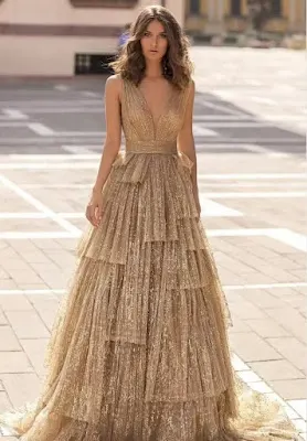 Shimmering Gold Tiered Evening Gown A model poses outdoors in a paved square, wearing a glamorous floor-length gold gown covered in fine glitter. The dress features a deep V-neckline, a defined waist, and a voluminous skirt crafted from multiple layers of pleated, shimmering fabric.