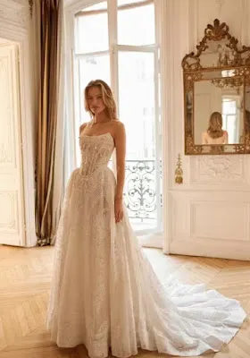 Elegant Corset-Style Lace Wedding Dress A woman standing in a luxurious, classic room wearing a white, strapless corset-style wedding dress featuring an intricate lace bodice and a full, flowing skirt with a long train.