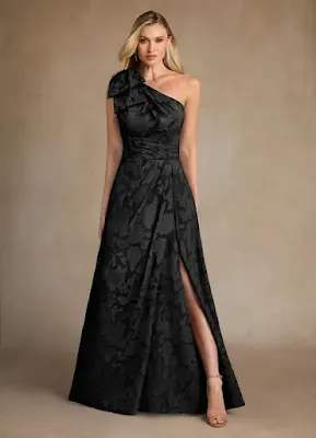 Black One-Shoulder Split Prom Dress Blonde model wearing a long black one-shoulder jacquard prom dress featuring a subtle floral pattern, a bow detail on the shoulder, and a high leg split.