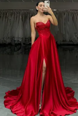 Uma mulher em pé, tirando uma foto no espelho, veste um longo vestido de baile vermelho de cetim. O vestido é sem alças, com um corpete espartilho estruturado e adornado com miçangas, uma saia ampla com cauda e uma fenda alta na perna. Ela usa saltos altos prateados e o fundo é uma loja de vestidos de noiva com araras de roupas desfocadas.