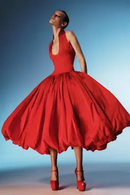 Uma modelo com cabelo curto e escuro posa dramaticamente, olhando para cima, em um fundo gradiente azul. Ela veste um vestido de baile vermelho vibrante, na altura do tornozelo, com corpete ajustado, decote em V profundo e uma saia bufante (bubble skirt) extremamente volumosa. Ela completa o visual com sapatos de salto plataforma vermelhos combinando.
