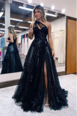 Long Black Prom Dress with Slit and Sparkle Blonde young woman posing in a fitting room wearing a long black prom dress with a one-shoulder design, shimmering tulle fabric, and a deep side slit.