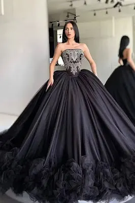 Vestido de Baile Preto com Corpete Bordado Uma mulher de cabelo preto comprido está em um provador e usa um vestido de baile preto extravagante com um corpete sem alças, detalhadamente bordado em prata e pedrarias, e uma saia de tule em camadas muito volumosa que termina com uma barra de babados. O reflexo dela é visível em um espelho ao fundo.