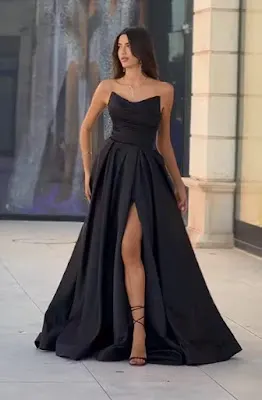 Black Evening Dress with Deep Slit and Sweetheart Neckline A tall woman with long brown hair poses outdoors wearing an elegant strapless black evening dress. The dress features a draped sweetheart neckline bodice and a full satin skirt with a high, bold leg slit. She completes the look with black strappy sandals and a delicate necklace. The background shows a store facade with a reflection.